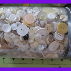 May include: A glass jar filled with a variety of white, cream, and yellow buttons. The jar has a silver lid.