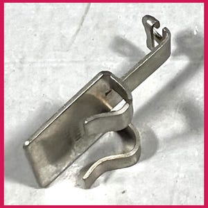 Pu&ograve; includere: Un piedino per macchina da cucire color argento. Il piedino ha una base rettangolare con un gancio curvo e un braccio angolato rialzato. Il metallo ha un aspetto leggermente usurato.