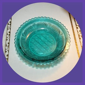May include: A teal-colored glass dish set with a scalloped edge. The dish features a detailed embossed pattern on both the lid and the base. The design includes floral and geometric elements, creating a vintage aesthetic. The set is circular and appears to be made of pressed glass.