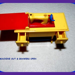 May include: A yellow toy sewing machine with a red top and two open drawers. The machine is on a white surface. The text "MACHINE OUT & DRAWERS OPEN" is visible below the machine.