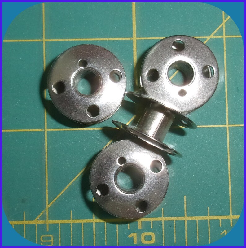 May include: Three silver metal bobbins for a sewing machine. The bobbins are round with a hole in the center and four smaller holes around the edge.