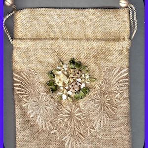 May include: A small, beige burlap drawstring bag with a white and green embroidered floral design. The bag has a simple, rustic look.