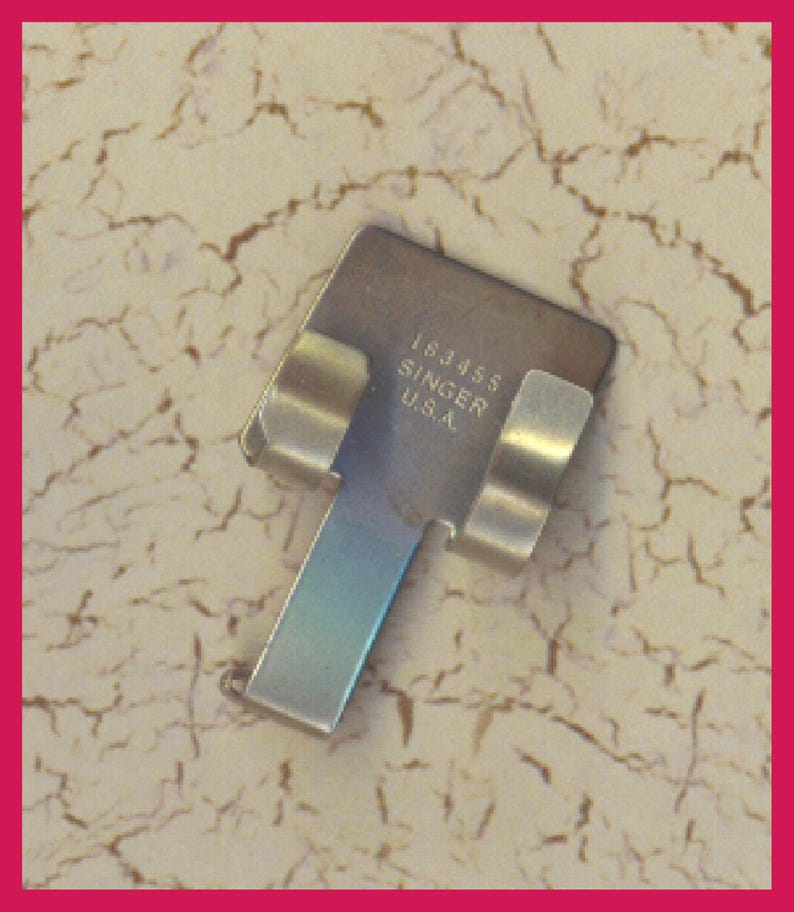 Pu&ograve; includere: Un accessorio per macchina da cucire Singer U.S.A., modello 163455, realizzato in metallo color argento. L'accessorio ha una parte superiore rettangolare con bordi curvi e una lunga, stretta estensione rettangolare. Due pezzi di metallo curvi si estendono dalla parte superiore.
