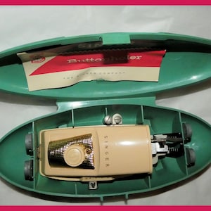 May include: An open, teal-colored case containing a vintage Singer buttonholer machine. The machine is beige with a gold-colored front and a round button. A red and white paper with the text "Buttonholer" is inside the case.
