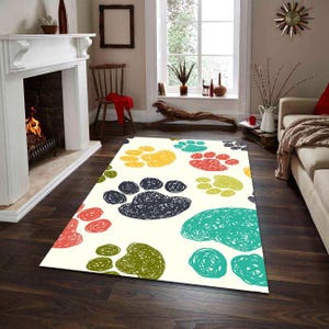 Pode incluir: Um tapete retangular com fundo creme e pegadas coloridas em amarelo, vermelho, verde, azul e preto. O tapete está colocado sobre um chão de madeira escura numa sala de estar.