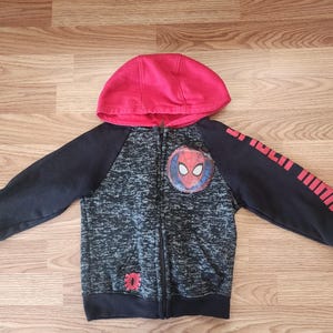 Puede incluir: Una sudadera con capucha con cremallera para ni&ntilde;os con capucha roja y mangas negras. La parte delantera tiene un patr&oacute;n moteado gris y negro con un emblema de Spider-Man. La manga derecha tiene rayas y texto rojos. La sudadera est&aacute; hecha de un material suave.