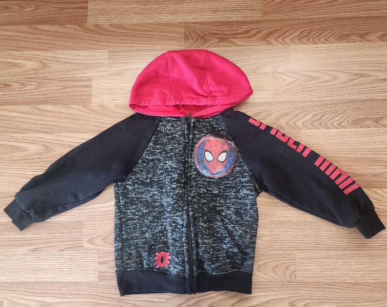 Puede incluir: Una sudadera con capucha con cremallera para ni&ntilde;os con capucha roja y mangas negras. La parte delantera tiene un patr&oacute;n moteado gris y negro con un emblema de Spider-Man. La manga derecha tiene rayas y texto rojos. La sudadera est&aacute; hecha de un material suave.