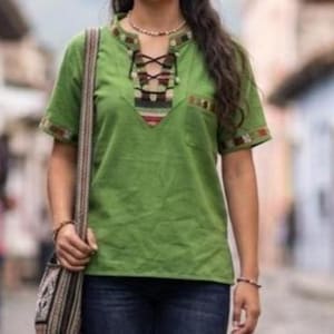 May include: A short-sleeved green shirt featuring a V-neckline with black lace-up detailing and a multi-colored striped pattern. The shirt includes a chest pocket and matching striped trim on the sleeve cuffs and neckline, paired with dark blue jeans.