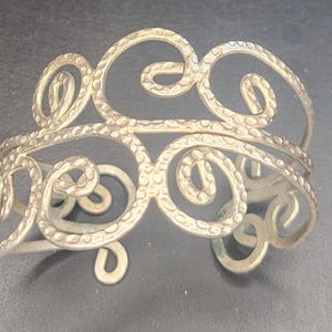 以下が含まれることがあります： シルバーカラーのカフブレスレット。オープンワークのデザインで、相互接続された渦巻き模様が特徴です。金属はテクスチャ加工された表面を持ち、ジュエリーに視覚的な魅力を加えています。