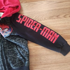 Puede incluir: Una sudadera con capucha con cremallera negra y gris con capucha roja. La manga derecha tiene las palabras "SPIDER-MAN" en letras rojas. Un emblema de Spider-Man est&aacute; en el pecho izquierdo.
