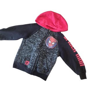 Puede incluir: Una sudadera con capucha con cremallera con el tema de Spider-Man. La capucha es roja y las mangas son negras con la inscripci&oacute;n "Spider-Man" en rojo. La parte delantera presenta un emblema de Spider-Man y un peque&ntilde;o logotipo de Spider-Man.