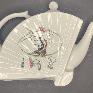May include: White ceramic teapot in a fan shape, featuring a painted illustration of a figure in traditional attire. The design includes a handle, spout, and a lid with a small knob. The illustration has red, green, and black accents.