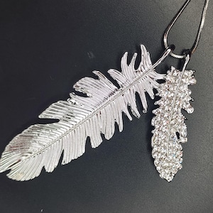 Może przedstawiać: Srebrny naszyjnik z zawieszką w kształcie pióra. Naszyjnik posiada dwie zawieszki w kształcie piór, jedno duże i teksturowane, drugie mniejsze i inkrustowane przezroczystymi kryształami. Wisi na cienkim, srebrnym łańcuszku, na czarnym tle.