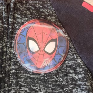 Puede incluir: Chaqueta con cremallera negra y gris con un gran emblema circular de Spider-Man en el pecho. El emblema presenta la cara del personaje en rojo y blanco, con un fondo azul de dise&ntilde;o de telara&ntilde;a. La chaqueta tiene una cremallera negra y una manga azul oscuro.
