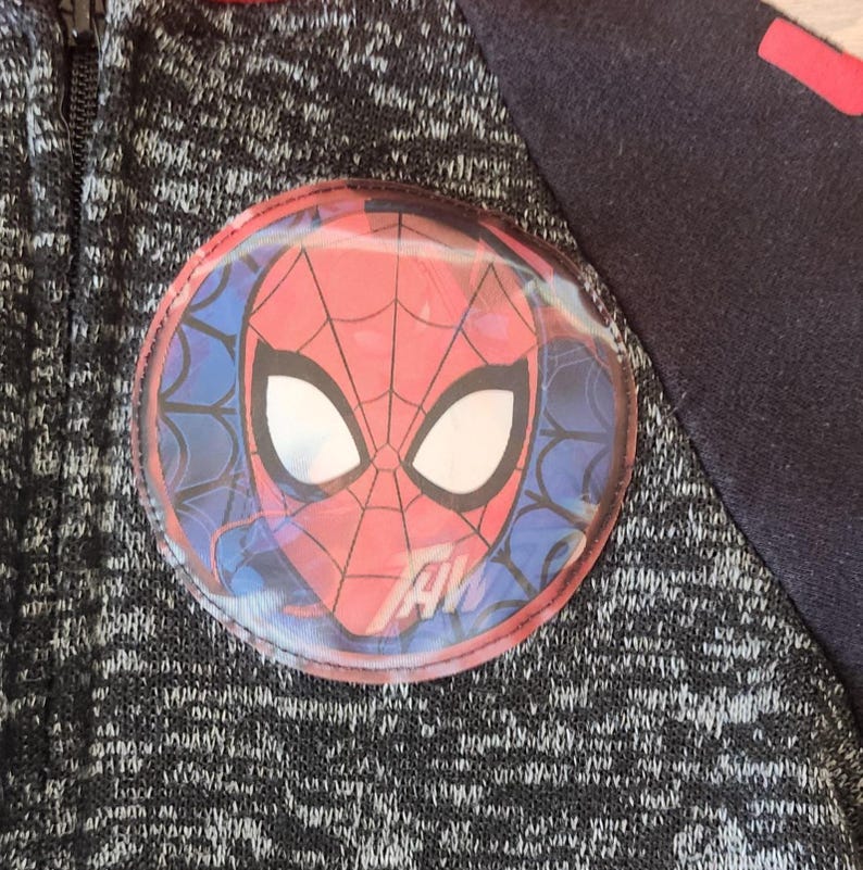 Puede incluir: Chaqueta con cremallera negra y gris con un gran emblema circular de Spider-Man en el pecho. El emblema presenta la cara del personaje en rojo y blanco, con un fondo azul de dise&ntilde;o de telara&ntilde;a. La chaqueta tiene una cremallera negra y una manga azul oscuro.