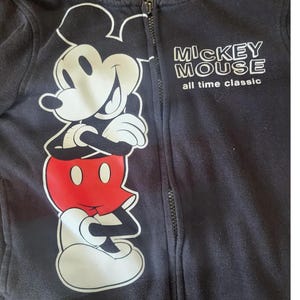 May include: Black zip-up hoodie featuring a large graphic of Mickey Mouse in white, red, and black. The text "MICKEY MOUSE all time classic" is printed in white on the right side of the hoodie. The hoodie has a full-length zipper.