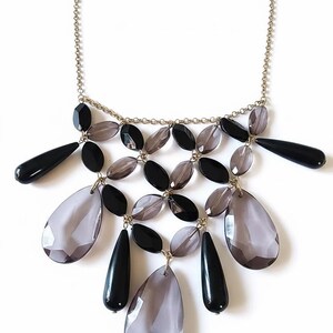 May include: A statement necklace featuring a gold-tone chain with a multi-layered bib design. The piece is adorned with alternating black and translucent grey teardrop-shaped beads, arranged in a geometric pattern that cascades downward.