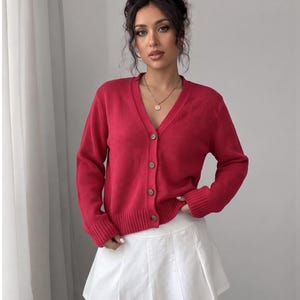 May include: A vibrant magenta long-sleeve cardigan featuring a V-neckline, a front button closure with four visible buttons, and ribbed cuffs and hem, paired with a solid white pleated mini skirt that has a clean, structured design.