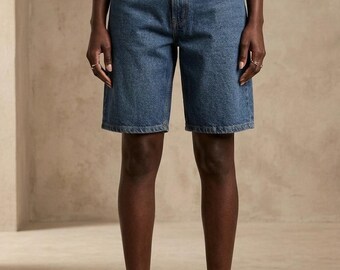 Puede incluir: Pantalones cortos de mezclilla azul medio con cintura alta, diseño clásico de cinco bolsillos y corte de pierna recta que llega por encima de la rodilla. Los pantalones se cierran con un botón metálico y cremallera, junto a un cinturón trenzado marrón.