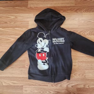 May include: Black zip-up hoodie featuring a large graphic of Mickey Mouse in white, red, and black. The hoodie has a hood and the words "Mickey Mouse all time classic" printed on the front. The garment is made of a soft material.