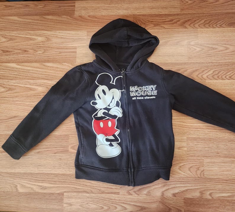 May include: Black zip-up hoodie featuring a large graphic of Mickey Mouse in white, red, and black. The hoodie has a hood and the words "Mickey Mouse all time classic" printed on the front. The garment is made of a soft material.
