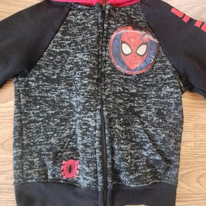 Puede incluir: Una sudadera con capucha con cremallera negra y gris con capucha y mangas rojas. La sudadera con capucha presenta un emblema de Spider-Man en el pecho y un gr&aacute;fico de ara&ntilde;a roja en la parte inferior izquierda. Las mangas tienen detalles en rojo.