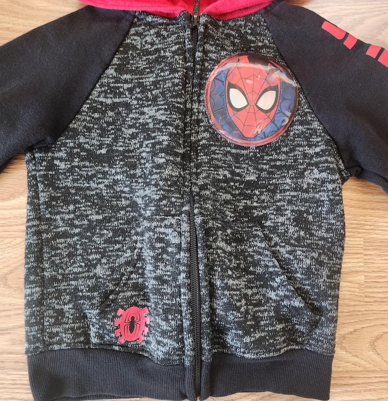 Puede incluir: Una sudadera con capucha con cremallera negra y gris con capucha y mangas rojas. La sudadera con capucha presenta un emblema de Spider-Man en el pecho y un gr&aacute;fico de ara&ntilde;a roja en la parte inferior izquierda. Las mangas tienen detalles en rojo.