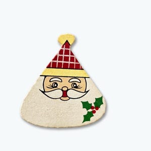 May include: A flat, triangular-shaped Santa Claus ornament featuring a red and white plaid hat with a yellow brim, a white beard, and a green holly leaf accent with three red berries on the lower right side against a cream-coloured background.