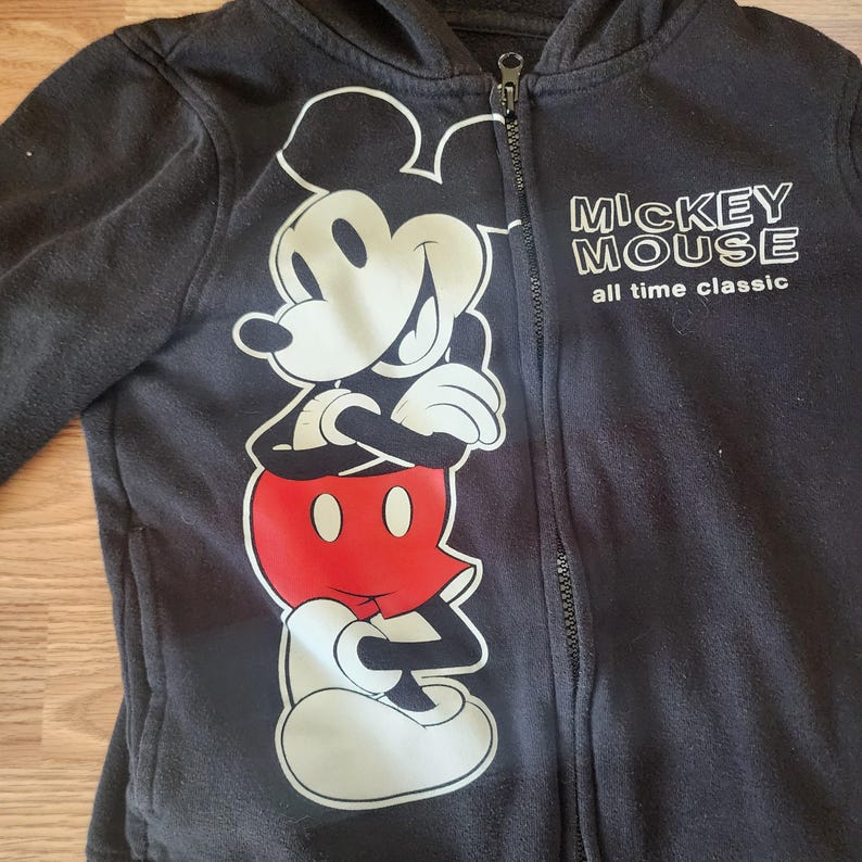 May include: Black zip-up hoodie featuring a large graphic of Mickey Mouse in white, red, and black. The hoodie has the text "MICKEY MOUSE all time classic" printed in white. The jacket has a full-length zipper and a hood.