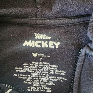 May include: Close-up of a navy blue Disney Junior Mickey Mouse hoodie label. The label features the text "Disney Junior MICKEY" in white, along with care instructions and size 7. The hoodie is made of 60% cotton and 40% polyester.