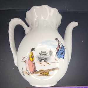 May include: White ceramic pitcher with a ruffled top, curved handle, and spout. The pitcher features a painted scene of two figures in traditional Korean clothing near a bridge and a wooden plank.
