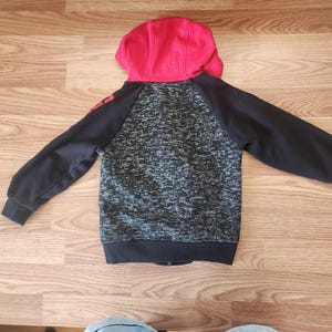 Puede incluir: Una sudadera con capucha negra y gris con capucha roja. La sudadera tiene mangas negras y un cuerpo gris y negro moteado. La palabra "RED" est&aacute; impresa en la manga izquierda. La sudadera est&aacute; hecha de un material suave y c&oacute;modo.