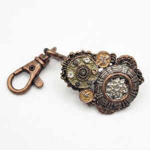 Può includere: Un portachiavi in metallo color rame con un moschettone attaccato a un gruppo di componenti circolari simili a ingranaggi. L'elemento decorativo presenta dischi metallici testurizzati, piccoli cristalli trasparenti e intricati motivi in rilievo.