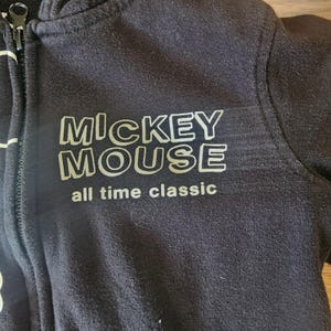 May include: Black zip-up hoodie featuring the text "MICKEY MOUSE all time classic" in white. The hoodie has a full zipper and a hood. The fabric appears to be a soft, dark material.
