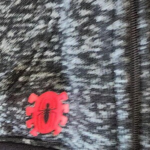 Puede incluir: Primer plano de una tela estampada gris y blanca con un emblema rojo con forma de insecto. El emblema presenta un contorno de insecto negro dentro de un marco rojo estilizado. La tela tiene un aspecto texturizado.