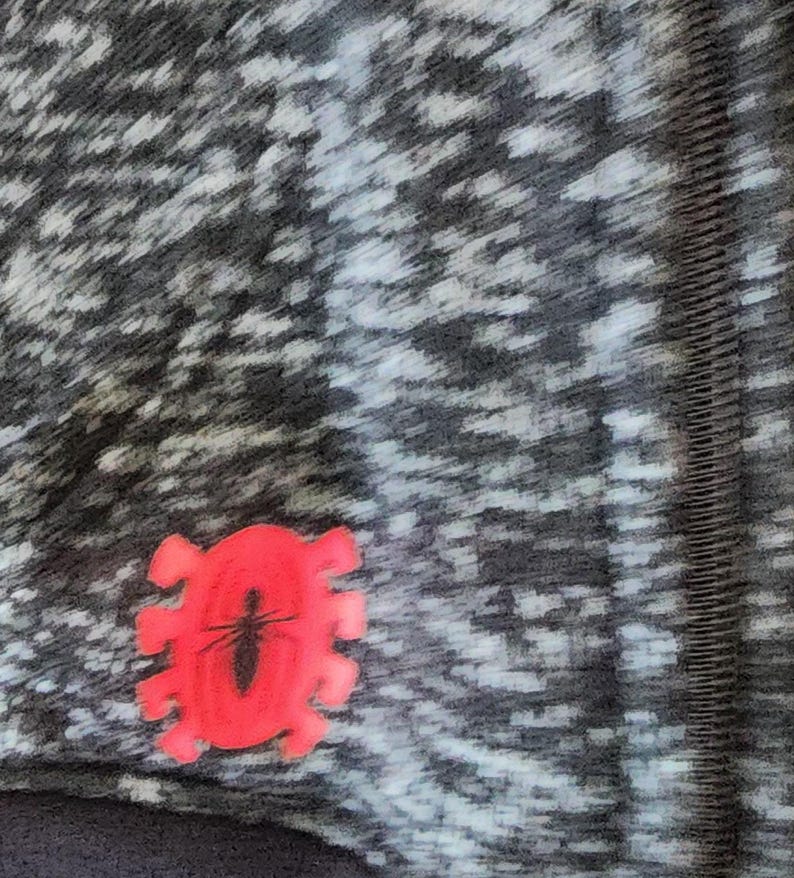 Puede incluir: Primer plano de una tela estampada gris y blanca con un emblema rojo con forma de insecto. El emblema presenta un contorno de insecto negro dentro de un marco rojo estilizado. La tela tiene un aspecto texturizado.