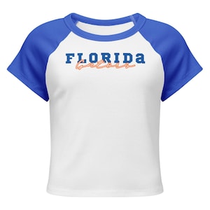 May include: White crop top with blue raglan sleeves and a blue crew neck. The shirt features the text "FLORIDA Gators" in blue and orange lettering across the chest. A sporty, casual top.
