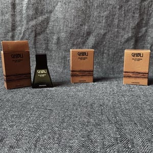 Può includere: Un assortimento di flaconi e scatole di profumo. Le scatole marroni hanno un testo nero che dice "GHIBLI". Un flacone di profumo nero è al centro. Lo sfondo è un tessuto grigio strutturato.