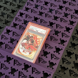 May include: A graded trading card in a plastic case, featuring a character illustration. It sits on a purple surface with a repeating skull and crossbones design. The background has a similar pattern in black.