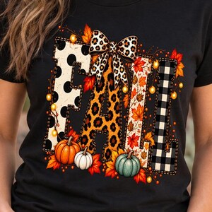 Peut inclure: Un t-shirt noir à manches courtes avec le mot FALL en grandes lettres majuscules. Chaque lettre présente un motif unique, notamment des pois noirs, un imprimé léopard, des feuilles d'automne et un motif écossais noir et blanc.