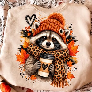 Peut inclure: Un sweat-shirt beige orné d'un motif de raton laveur portant un bonnet en tricot orange et une écharpe à imprimé léopard. Le raton laveur tient un gobelet à café avec un cœur, entouré de feuilles d'érable orange, de cœurs et de points noirs.