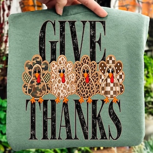 Peut inclure: Un sweat-shirt vert sauge avec le texte noir GIVE THANKS. Entre les mots se trouvent quatre dindes aux motifs variés sur leurs plumes : camouflage, floral, imprimé léopard et un motif à carreaux marron et blanc.