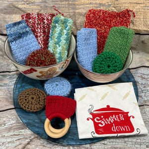 Peut inclure: Deux bols en métal remplis d'articles de cuisine crochetés colorés, dont des débarbouillettes et des éponges. Une assiette en verre bleu contient des articles supplémentaires, dont un anneau de dentition rouge et un torchon blanc avec un dessin de pot rouge et les mots "Simmer down."