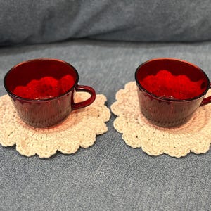 May include: Two ruby red glass teacups with handles rest on cream-colored, crocheted doilies. The cups have a textured surface. The doilies have a scalloped edge. The cups and doilies are on a blue denim surface.