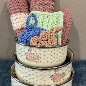 May include: A stack of three cream-colored enamel bowls with floral designs, filled with various crocheted items in shades of pink, blue, and green. The bowls have silver rims and are stacked on top of each other.