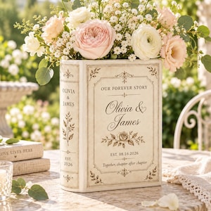 Puede incluir: Un florero de cerámica en forma de libro de color crema con el texto "OUR FOREVER STORY," "Olivia & James," "EST. 08.16.2026," y "Together, chapter after chapter." El florero está lleno de un ramo de rosas rosas y blancas y follaje.