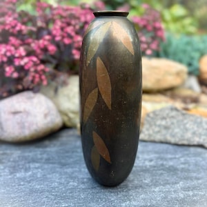 Puede incluir: Un jarrón cilíndrico de metal con un acabado en tono bronce oscuro, que presenta un patrón repetitivo de formas de hojas incrustadas de color más claro. El jarrón tiene una abertura circular estrecha y un cuerpo liso y redondeado.