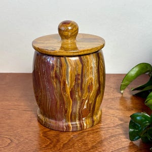 May include: A cylindrical stone jar with a matching lid featuring a spherical knob. The jar is crafted from polished stone displaying vertical bands of deep burgundy, golden yellow, and brown hues throughout the entire exterior surface.