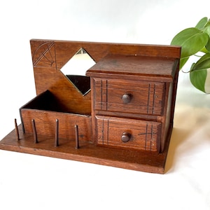 May include: A brown wooden desk organizer featuring two stacked drawers with round knobs, an open storage compartment, and four vertical pegs on the base. A small diamond-shaped mirror is set into the back panel, which also displays a carved fan-like design in the upper left corner.
