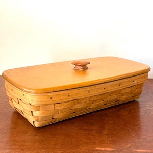Può includere: Un cestino rettangolare in legno intrecciato con un coperchio liscio in legno marrone chiaro. Il coperchio presenta un piccolo pomello quadrato centrale. Il corpo del cestino è realizzato con sottili strisce di legno intrecciate con chiodini metallici scuri.
